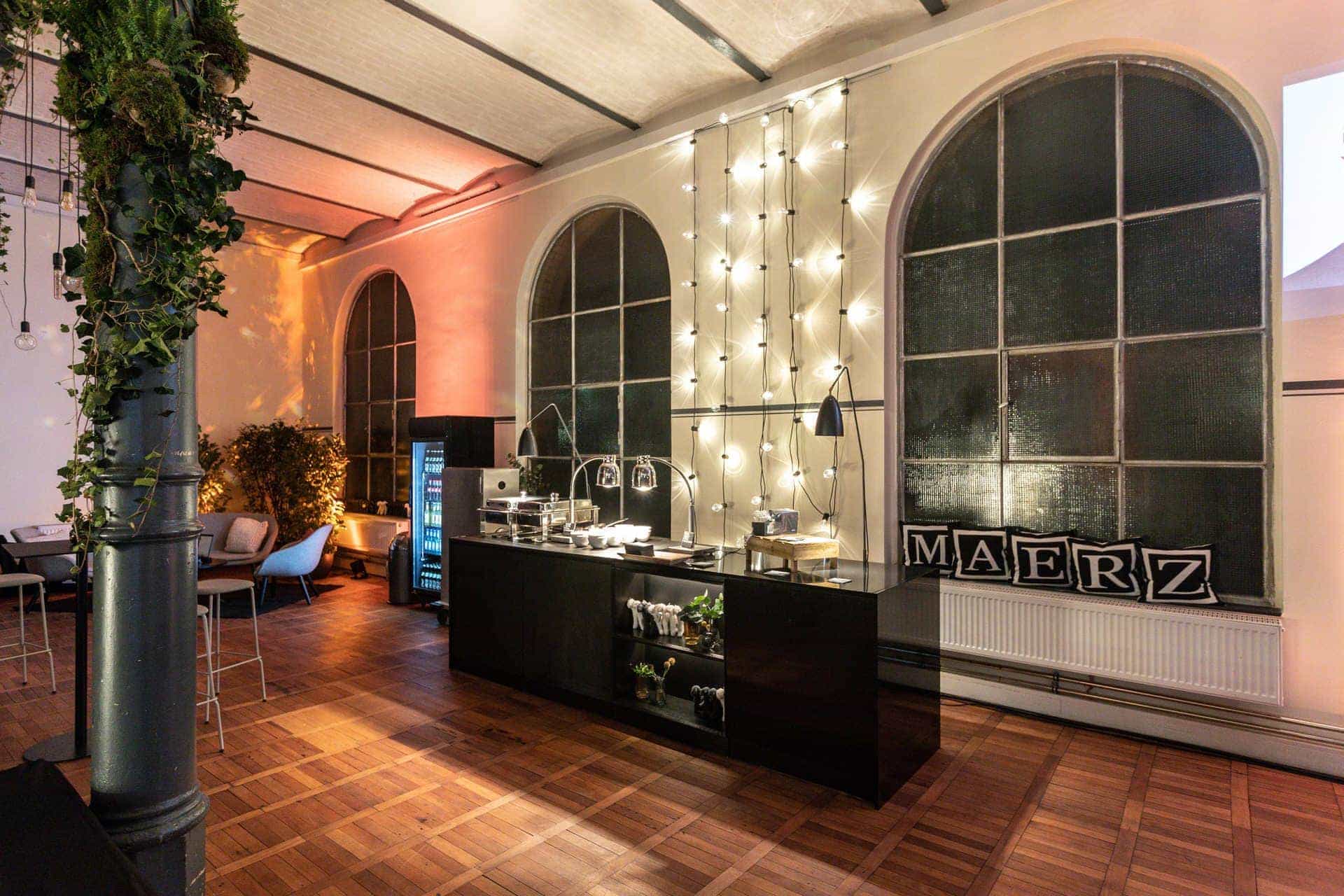 Stilvolle Event-Ecke im Magazin in der Heeresbäckerei, dekoriert mit Lichterketten und Pflanzen. Ein eleganter schwarzer Tresen mit Speisen und Getränken, daneben Sitzgelegenheiten und dekorative Kissen.