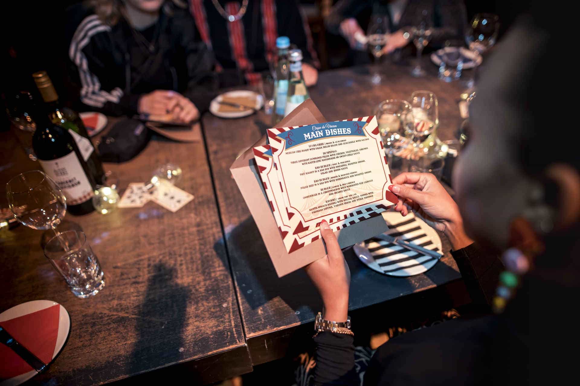 Ein Gast hält eine Menükarte bei einem Event von PENG Communications GmbH im Magazin in der Heeresbäckerei. Auf dem Tisch stehen Weinflaschen, Gläser und dekorative Kerzen